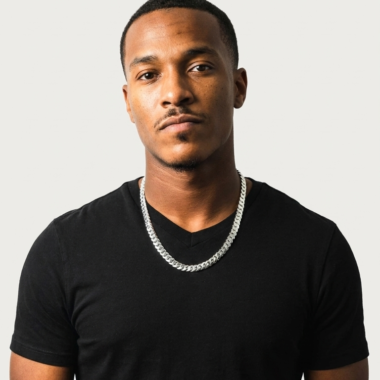 A man wearing a black v-neck t-shirt and a thick silver chain necklace, photographed against a light gray studio background.